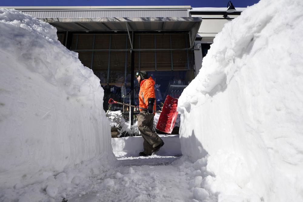 A Cohasset, Mass., resident carries a snow shovel past a snow bank on a sidewalk, Sunday, Jan. 30, 2022, in Scituate, Mass. Gusty winds and falling temperatures plunged the East Coast into a deep freeze as people dig out from a powerful nor’easter that dumped mounds of snow, flooded coastlines and knocked power out. (AP Photo/Steven Senne)