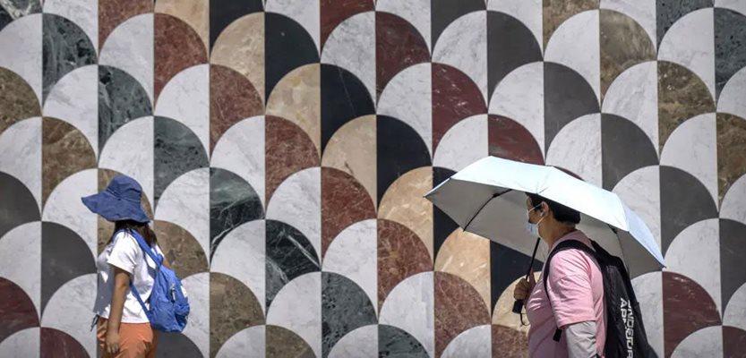 People use a sun hat and an umbrella to protect themselves from the sun as they walk outside a shopping mall on an unseasonably hot day in Beijing, Saturday, June 24, 2023. (AP Photo/Mark Schiefelbein)